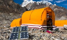 an orange tent with two solar panels on the ground in front of some rocks and snow capped mountains