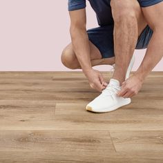 a man tying his white tennis shoes on top of a wooden floor in front of a pink wall