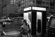 a woman standing next to a phone booth on a city street with an umbrella over her head