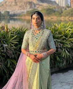 a woman in a green and yellow sari standing next to some bushes with buildings in the background