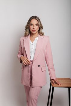 a woman standing next to a table wearing a pink suit and white shirt with her hands in her pockets