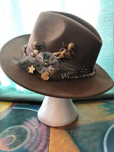 "Very unique soft brown felt fedora hat, \"Indiana Jones\" style by and hand decorated by Abracadebora in a \"Time is Precious so Hold on to your Hat\" theme. This is a one of a kind art creation and definitely a conversation piece. This soft brown fedora carries cupids, and hearts, a clock piece and feathers set into a vintage beaded brown ribbon band as a reminder that time is precious and cherished. Wearable art and unusual treasure." Unique Brown Hat With Curved Brim, Handmade Brown Felt Hat With Flat Brim, Brown Handmade Mini Hats With Curved Brim, Handmade Brown Mini Hats With Curved Brim, Handmade Brown Mini Hat With Short Brim, Vintage Handmade Brown Hat Bands, Vintage Brown Handmade Hat Bands, Unique Brown Fedora With Flat Brim, Unique Brown Fedora With Short Brim