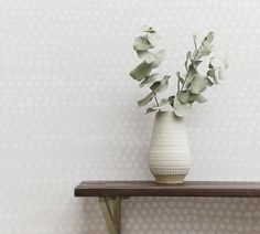 a white vase filled with green leaves on top of a wooden table next to a wall