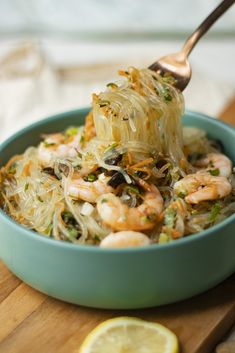 a bowl filled with noodles and shrimp on top of a wooden cutting board next to a lemon wedge