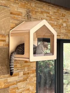 a cat sitting on top of a wooden house shaped like a birdhouse hanging from the side of a brick wall