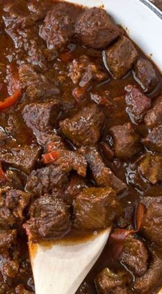 a white bowl filled with beef and carrots next to a wooden spoon on top of a table