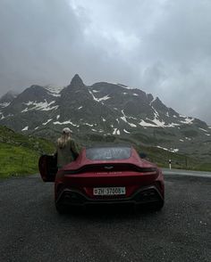 a person standing next to a red sports car in front of a mountain with snow on it