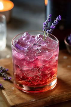 a glass filled with ice and purple flowers on top of a wooden cutting board next to a candle