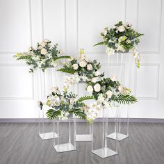 four clear vases with white flowers and greenery in them on a wooden floor