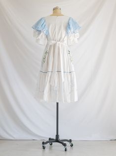 Experience garden-inspired elegance with our Sugar Cream Vintage Midi Dress. Made from upcycled cotton, it features a square neck, playful blue fabric ruffles on half cuff sleeves, and intricate blue daisy embroidery with lotus patchwork. Complete with an adjustable bow tie and tiered flared hemline, this dress exudes eco-chic charm! Care: Cool machine wash. Dry at a lower heat and press with a medium iron.  Condition: Embrace the distinctive charm of this upcycled treasure, where imperfections are unique features. Delight in the occasional loose thread or subtle discoloration, each adding to the character and history of this special garment. Broderie Anglaise Square Neck Dress For Garden Party, Cotton Square Neck Dress For Garden Party, Cotton Dresses For Garden Party With Square Neck, Spring Cotton Vintage Dress With Square Neck, Spring Square Neck Vintage Cotton Dress, Spring Cotton Prairie Dress With Square Neck, Blue Cotton Prairie Dress For Summer, Cotton Prairie Dress With Ruffles For Garden Party, Spring Square Neck Prairie Dress In Cotton