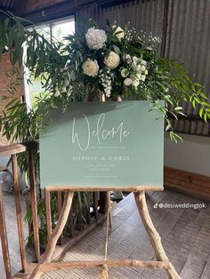 a welcome sign sitting on top of a wooden stand next to flowers and greenery