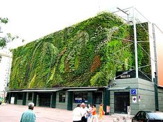 people are walking past a building covered in green plants on the side of the street