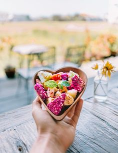 a person holding a heart shaped bowl filled with fruit and veggies on top of a wooden table