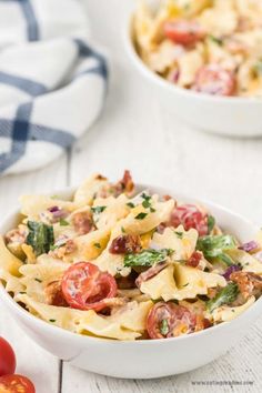 two bowls filled with pasta and vegetables on top of a white wooden table next to tomatoes