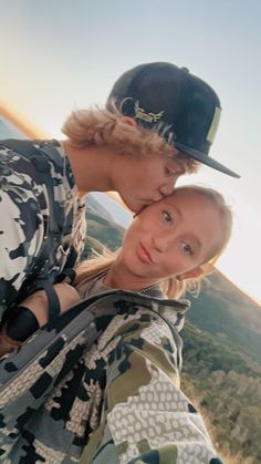 two people standing next to each other with mountains in the background and one person wearing a hat