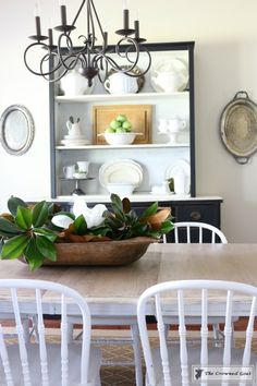 a dining room table with white chairs and a wooden bowl filled with flowers on it