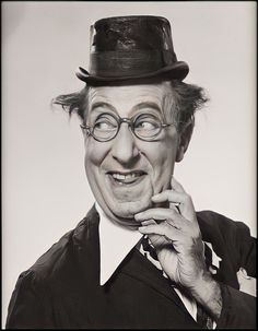 an old man wearing glasses and a top hat with his hand on his chin, smiling at the camera