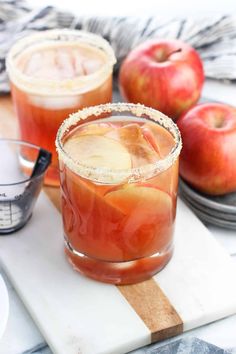 two glasses filled with apple cider sitting on top of a cutting board next to apples