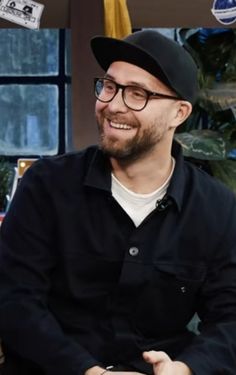 a man wearing glasses and a hat sitting in front of a window smiling at the camera