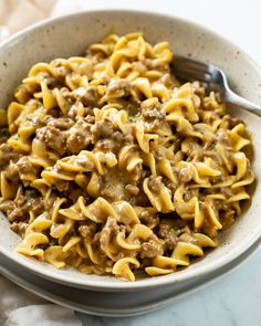 a white bowl filled with pasta and meat on top of a marble table next to a fork