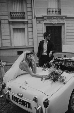 a woman in a wedding dress sitting on top of a white car next to a man