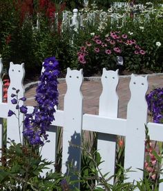 a white picket fence with purple flowers in the foreground and an orange cat statue behind it