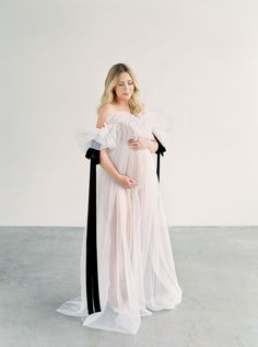 a pregnant woman wearing a white gown and black ribbon around her waist, standing in an empty room