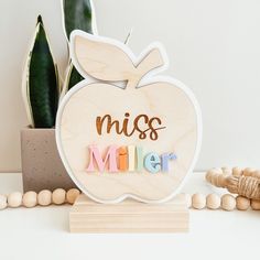 an apple shaped wooden sign with the word miss miller on it next to beads and a potted plant