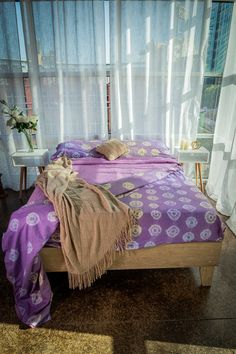 a bed with purple bedspread and pillows in front of a curtained window