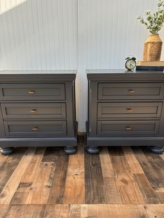 two gray nightstands side by side against a white wall and wooden floor with a clock on top