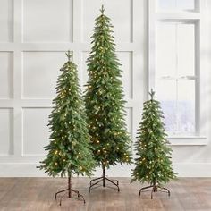 three artificial christmas trees with lights on them in front of a white wall and wooden floor