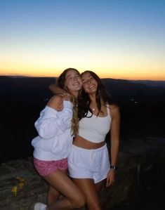 two young women hugging each other while standing on top of a mountain at sunset or dawn