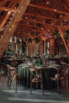 the inside of a barn with tables and chairs covered in green cloths, hanging lights and greenery