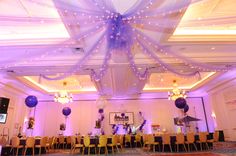 a banquet hall decorated with purple and white balloons, lights and drapes on the ceiling