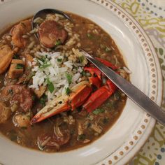 a white bowl filled with stew and meats on top of a tablecloth next to a spoon