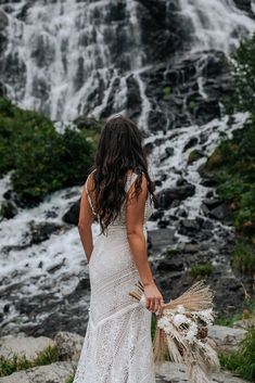 a woman standing in front of a waterfall wearing a wedding dress and holding a bouquet