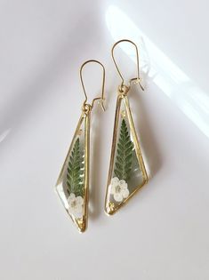 a pair of earrings with green leaves and white flowers in them on a white surface