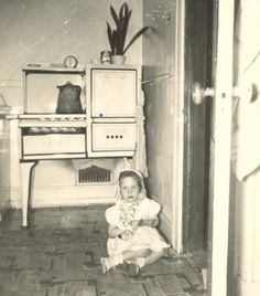 an old black and white photo of a baby sitting on the floor in front of a stove