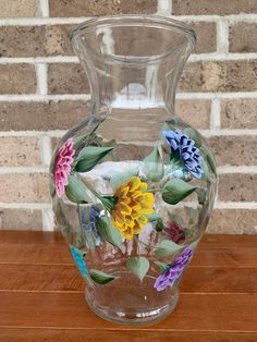 a glass vase with colorful flowers painted on it sitting on a wooden table next to a brick wall