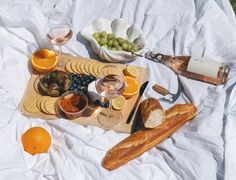 an assortment of food and wine on a white blanket with grapes, bread, oranges, and other foods