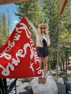 a woman holding up a red and white flag on top of a rock in front of trees