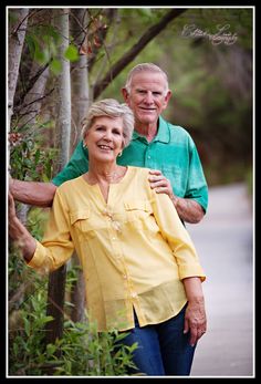 an older man and woman standing next to each other in front of trees with leaves on them