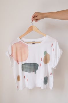 "Cropped organic cotton T-shirt, with round neck, white base and hand painted motifs in earth tones (dust pink, sand, gray green, gray, terracotta). This shirt is made in Africa with ethical craftsmanship. The cropped shape with rolled sleeves, combined with the manual water stains, make it perfect for both relaxed and sophisticated looks. You'll look radiant with a high-waisted jean or with dress pants and a jacket, achieving the \"effortless elegance\" effect that we love at APPA Studio. The \ White Hand Printed Crew Neck T-shirt, Conscious White Crew Neck Top, White Conscious Crew Neck Top, White Hand Painted Top For Summer, Hand Printed Organic Cotton T-shirt, Short Sleeve Cotton T-shirt With Natural Dye, White Crew Neck Top, Hand Printed Graphic Tee For Summer, Artsy White Short Sleeve T-shirt