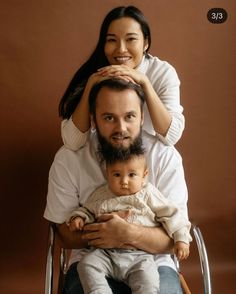 a man sitting in a chair holding a baby and smiling at the camera with his mother standing behind him