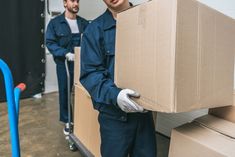 two men in blue work clothes are carrying cardboard boxes on a hand truck and one man is wearing white gloves