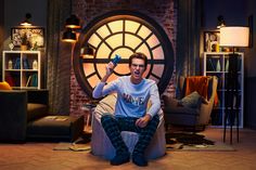 a man sitting on top of a couch in front of a large round window holding a remote control