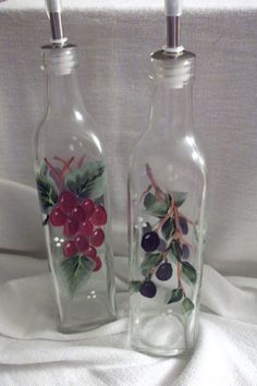 two glass bottles with fruit painted on them sitting on a white cloth covered tablecloth