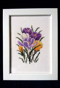 a white frame holding a cross stitch picture with purple and yellow flowers in it on a black background