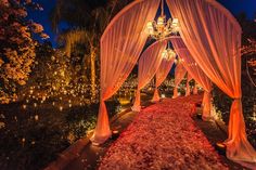 an outdoor wedding venue with white drapes and pink flowers on the ground at night