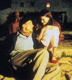 an old man sitting on the ground next to a young woman with her arm around him